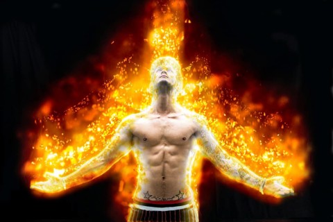 Tattooed muscular man with nude torso holding hands apart and burning in fire on black background