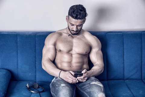 Handsome shirtless muscular bodybuilder man typing or surfing the internet with cell phone while