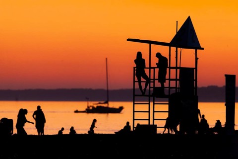 Silhouetted figures against an orange sunset on the beach with a lifeguard tower and a calm Lake