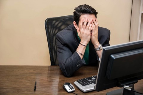 Desperate, worried young businessman sitting at his desk in front of his computer with his face in