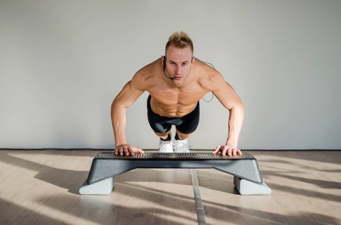 Young aerobics male coach shirtless leaning on step teaching class, looking at camera