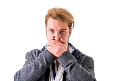 Young blond man covering his mouth while looking at camera against of white background