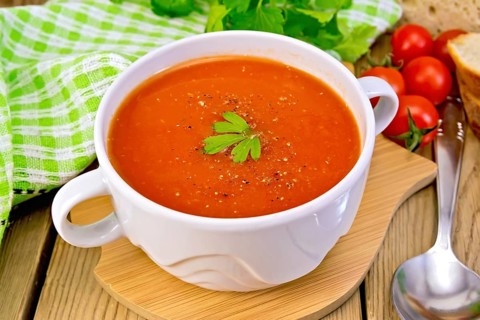 Tomato soup in a white bowl, green napkin, spoon, tomato, parsley, bread on a wooden boards