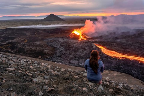 A woman sits on a mountain and looks at a glowing volcano, Grindavik, Sudurnes, Iceland, Europe