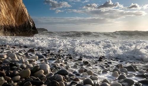 Low angle view of seascape with windy waves splashing on the rock and the sea pebbles. Rock of