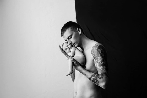 Father with tattoo on his arm is holding his newborn baby on a black and white background.