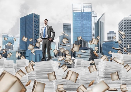 Confident businessman in suit standing on pile of documents among flying books with cityscape on