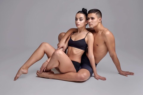A pair of a graceful athletic ballet dancers in black suits are posing over a gray studio