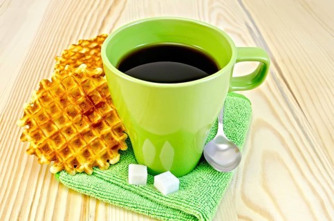 Two golden round waffles, green mug, sugar, spoon on green napkin on a background of wooden board