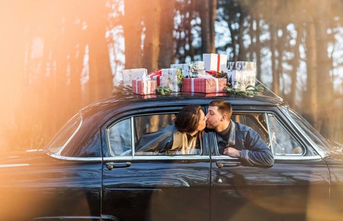 Loving man and woman sticking out of retro car windows and kissing with Christmas gifts on roof in