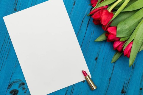 Red tulips, lipstick and a white sheet of paper for your greetings on the background of blue wooden