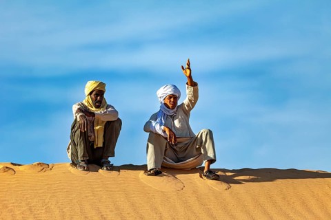 Two men sit on a sand hill in the desert under a clear blue sky, two Tuareg sit on a sand dune and