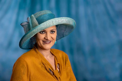 Woman wearing blue hat and yellow clothes, smiling in front of blue background, staged portrait