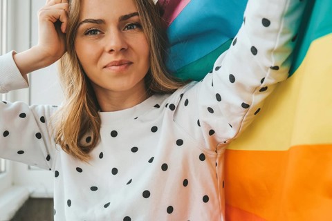 Young blonde millennial hippie woman with rainbow LGBTQ flag at home balcony. Peace and freedom the