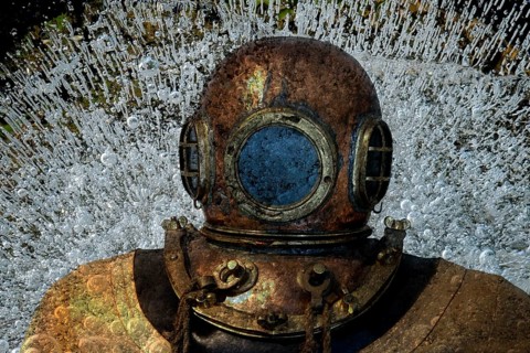 Metal diving helmet with round viewing windows amidst swirling air bubbles. Landscape format.