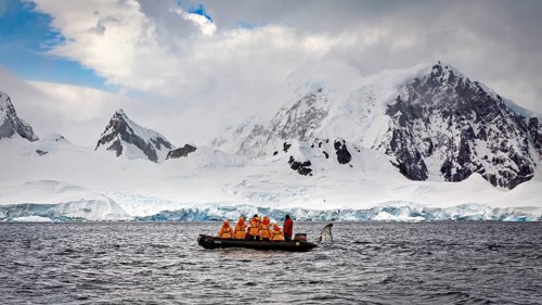 Group in a boat in the foreground, surrounded by snow-covered mountains and overcast sky, people in