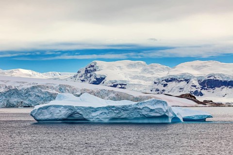 A large iceberg floats in the sea, surrounded by snowy mountains under a blue sky, iceberg in the