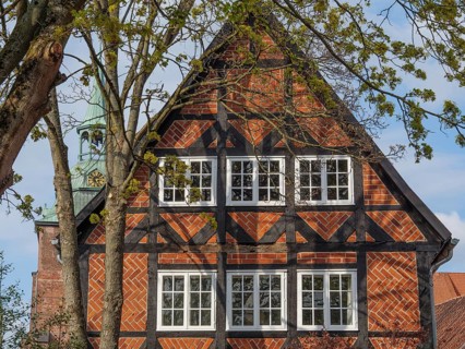 The picture shows a half-timbered house with red bricks, surrounded by trees. A church spire is