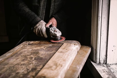 Close-up of hands sanding a wooden door in a dimly lit workshop next to a window