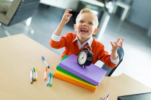 Cute blonde caucasian kid sit at the table with colorful folders, hold alarm clock, evil laugh,