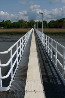 A long jetty stretches straight across the water, flanked by white railings under a blue sky,