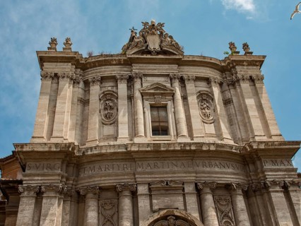 Front façade of a baroque building with intricate ornamentation and Latin inscription, framed in a