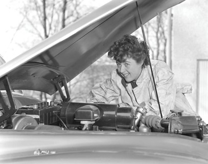 In the 1950s. A woman in an overall fixing something on the car that has it's hood open. Sweden