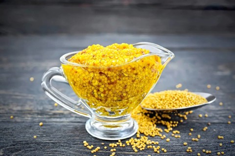 Mustard Dijon sauce in a glass sauceboat and mustard seeds in a spoon on a black wooden board