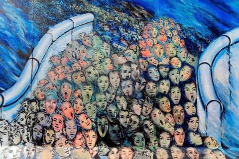 A multitude of surreal painted faces crowd together, surrounded by blue and bright colours,