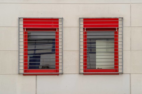 Two windows with red frames and metal grilles restrict the view outside