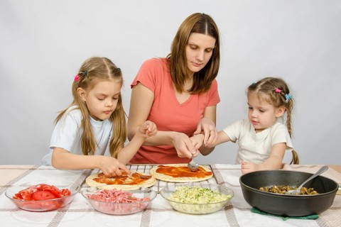 Mom helps younger daughter spread ketchup on a pizza, the eldest daughter, she is preparing a