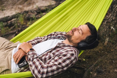 Man on bicycle trip at camping by lake is relaxing in green hammock while listening to music.