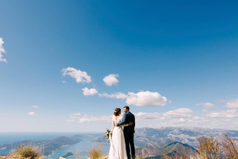 Newlyweds stand with their backs hugged against the backdrop of mountains and a bright blue sky.