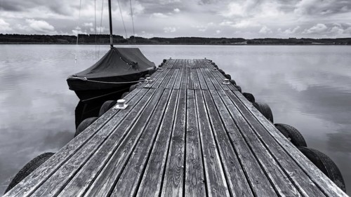 Wooden jetty and boat on a calm lake under a cloudy sky in black and white, Irsingener See, barrage