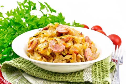 Cabbage stew with sausages in a white plate on a towel, tomatoes, parsley and a fork on the