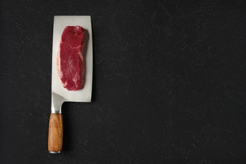 A piece of raw beef meat on a sharp cleaver with a wooden handle. The cleaver rests on a black
