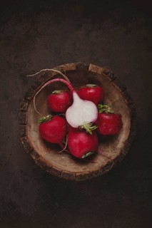 A collection of vibrant red radishes sits in a rustic wooden bowl. The contrasting dark background