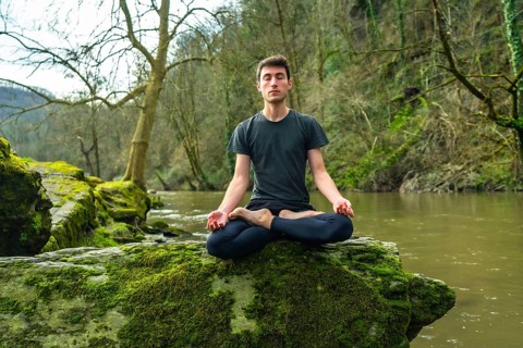 Yoga instructor meditating in lotus position on a mossy rock by a tranquil river, surrounded by