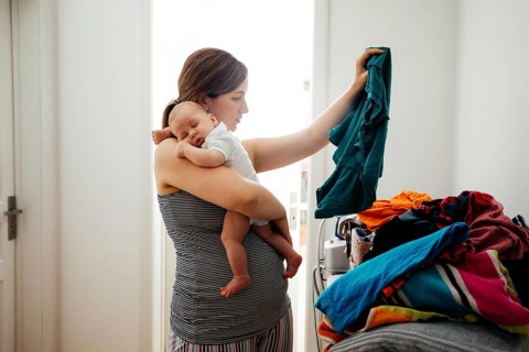 Young mother holding sleepeing baby on her shoulder and trying to sort the laundry simultaniously.