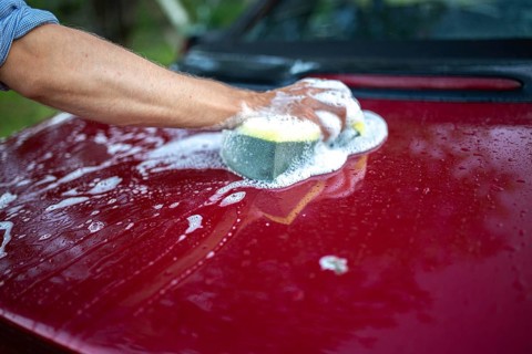 A person cleaning a red car with a sponge, creating soapy suds