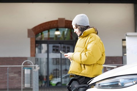 Woman with mobile phone in yellow jacket at charging station with electric car, environmentally
