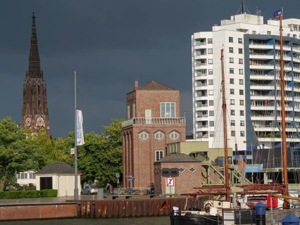 A cityscape with buildings, a church tower and ships in the harbour under a cloudy sky,