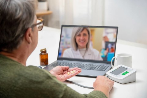 Senior woman having online medical consultation with doctor using laptop, holding pills in hand and