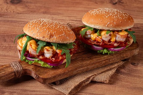 Two shrimp burgers and buns with sesame seeds, bacon, lettuce, tomatoes and onions, on a wooden
