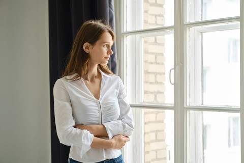 A teenager stands by a window, gazing outside with a thoughtful expression. The cozy room features
