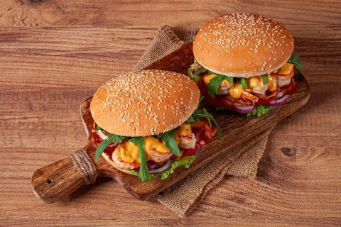 Two shrimp burgers and buns with sesame seeds, bacon, lettuce, tomatoes and onions, on a wooden