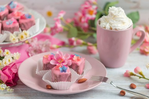 Pink petit fours with flower decoration on a plate next to a cup of coffee with cream