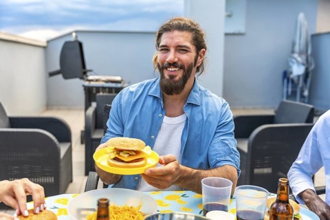 Happy man holding a burger during a rooftop barbecue party with friends, enjoying good food and