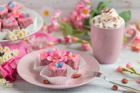 Pink petit fours with flower decoration on a plate next to a cup of coffee with cream