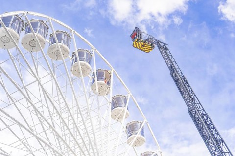 A crane near a Ferris wheel lifts equipment against a cloudy sky, fire brigade exercise for gondola